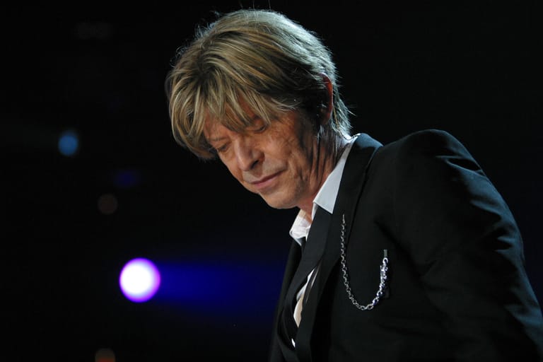 Performer in black suit and chain necklace under stage spotlight, head bowed.