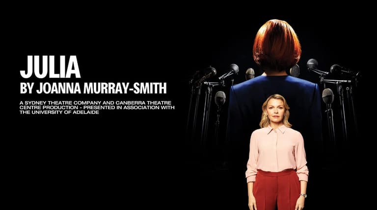 A woman in a pale pink shirt and red trousers stands before a large projected image of herself seated in a chair against a black background.
