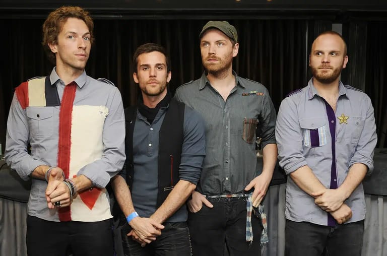 Four male band members stand in a line against a dark curtained backdrop, dressed in casual shirts and jackets.