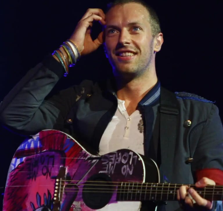 Musician in dark jacket holding a decorated acoustic guitar, hand to head, smiling under stage lighting.