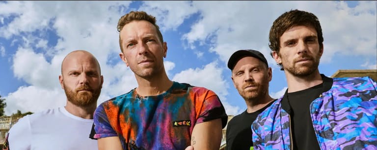 Four men stand outdoors against a blue sky, wearing colourful graphic clothing and patterned shirts.