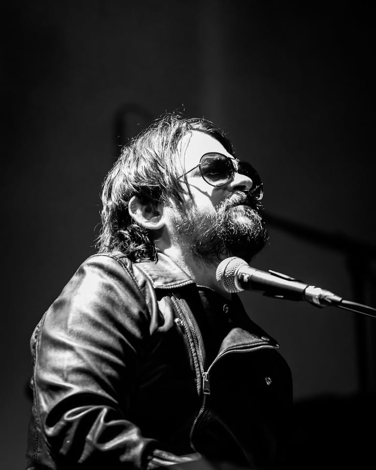Bearded musician wearing sunglasses and leather jacket performs at microphone in black and white.