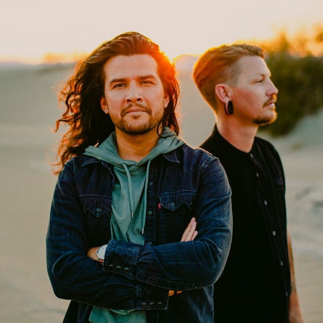 Two men with long hair stand outdoors at golden hour, one facing the camera with arms crossed, the other in profile.