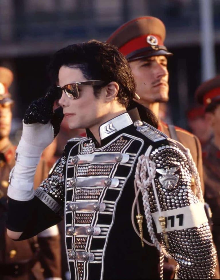 Person in sunglasses and ornate black studded jacket with white gloves, hand raised to face.