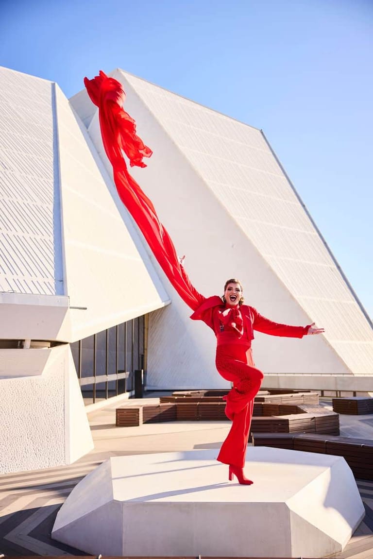 Performer in red attire striking a dramatic pose outside a modern white architectural building with flowing red fabric.