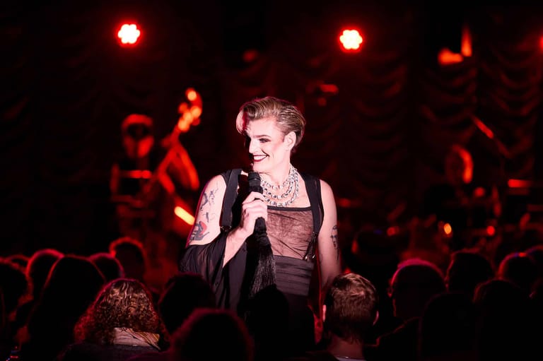 Performer singing into microphone on stage beneath red spotlights with band and crowd visible behind.