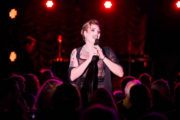 Singer holding microphone on stage amid red stage lighting and audience silhouettes.