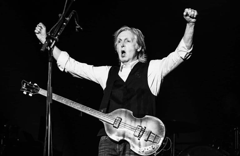 Musician with bass guitar performing at microphone with arms raised in celebration against black background.