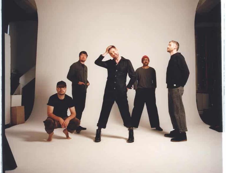 Five musicians in dark clothing posed in a bright studio setting with dramatic side lighting.