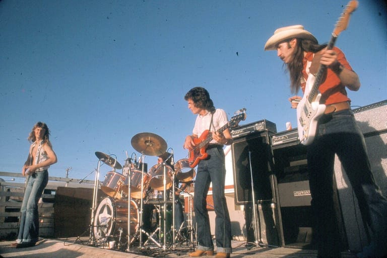 Live band performing outdoors in bright daylight, with guitarist and drummer visible on stage.