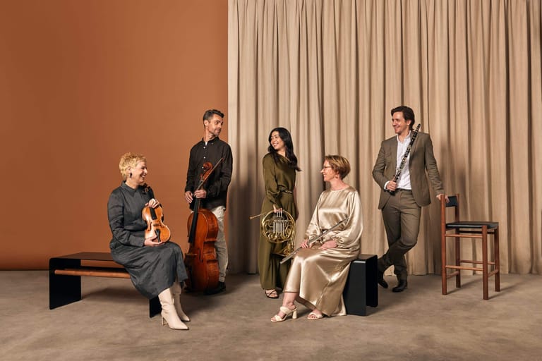 Five musicians with string instruments stand and sit against a warm ochre backdrop with draped curtains.