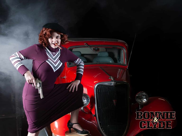 Performer in purple suit posing with handgun against a vintage red car in dramatic stage lighting.