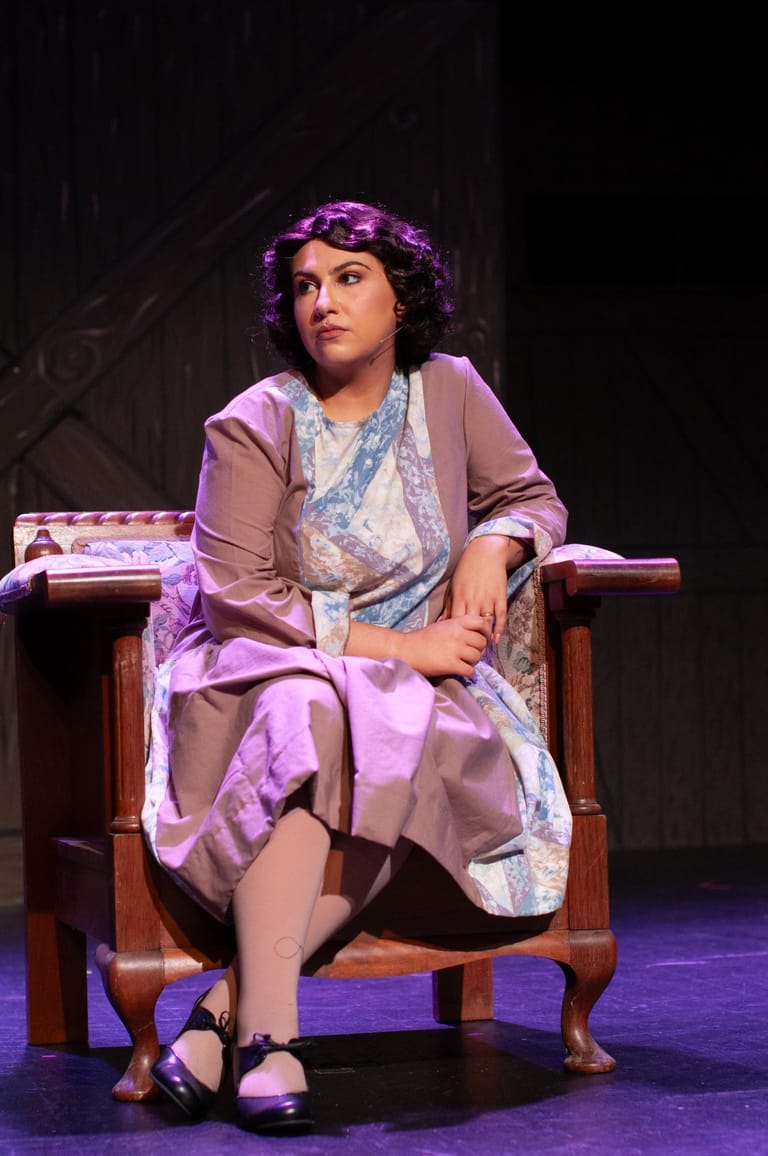 Actress seated on wooden chair in period costume, lit by purple stage lighting during Bonnie & Clyde theatre production.