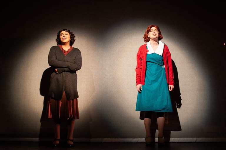 Two female performers stand under dramatic stage spotlights during a musical theatre production scene.