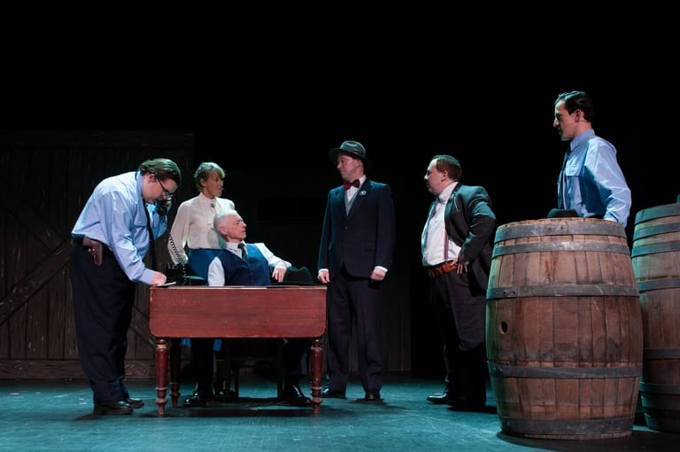 Performers in period costume gather around a wooden table on stage with barrels, lit by teal stage lighting during a theatrical production.