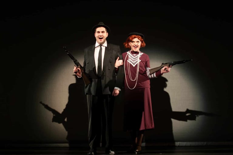 Two performers in 1930s gangster attire hold weapons on stage, centre-lit against a dark backdrop.