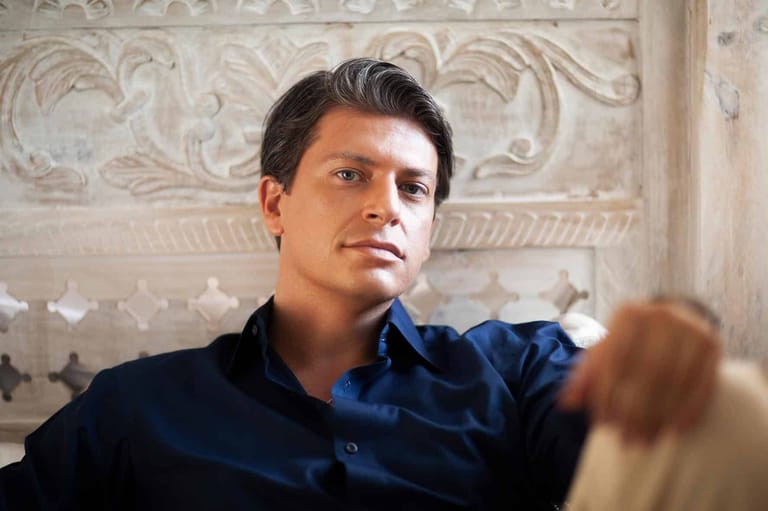 Man in dark blue shirt speaks indoors with ornate white wall panelling behind him.