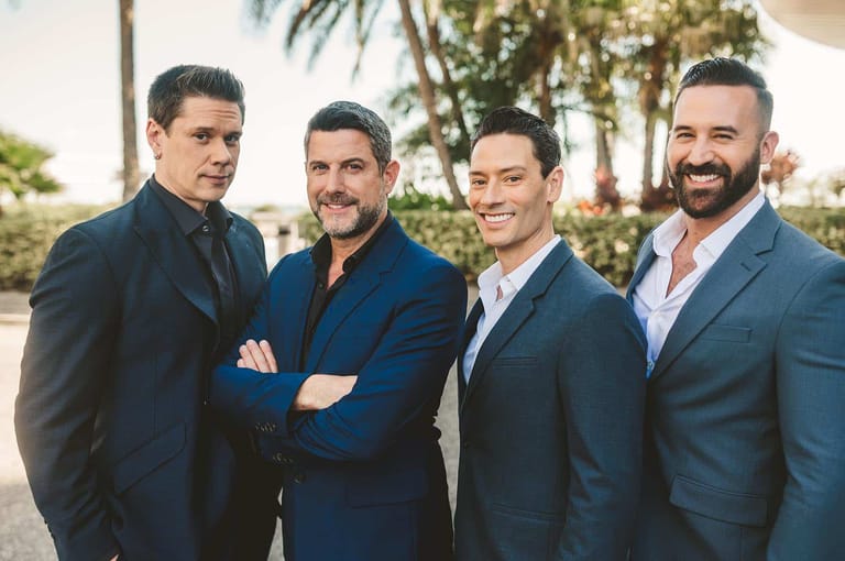 Four men in dark suits stand together outdoors with tropical plants in the background.