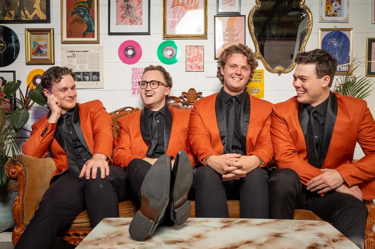 Four musicians in matching orange blazers sit together in a colourful art-filled room.