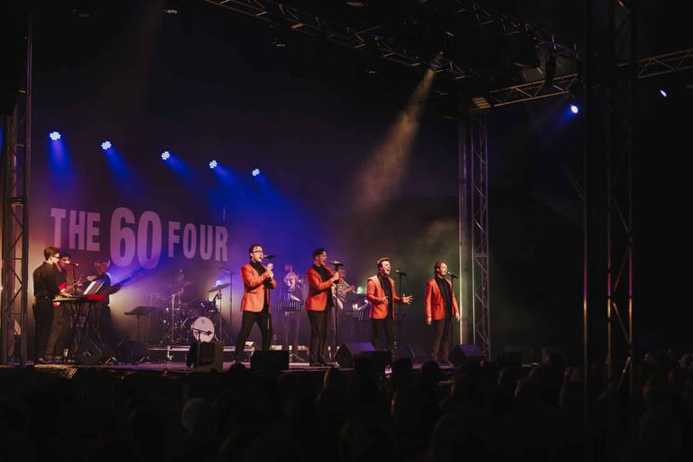 A cappella group performs on stage under blue spotlights with "The Go Four" signage visible behind them.