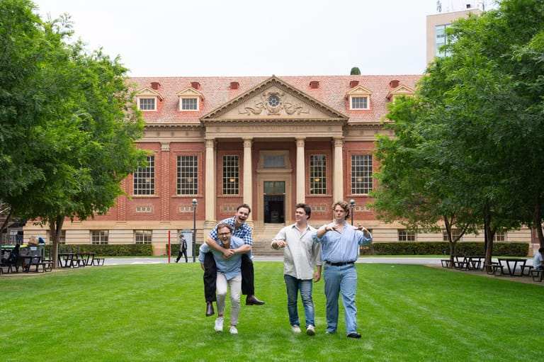 Four people stand on a manicured lawn facing a grand red-brick heritage building with classical architecture.
