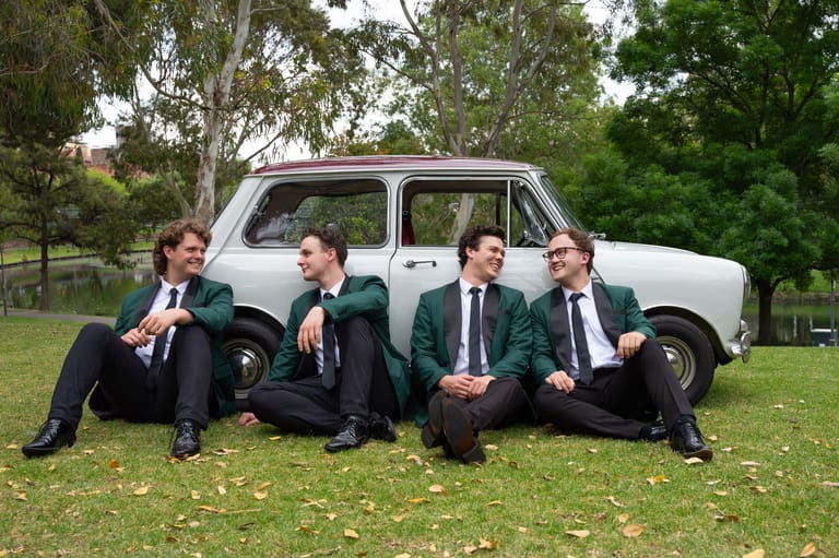 Four performers in school uniforms and blazers sit against a white vintage car in a grassy park setting.