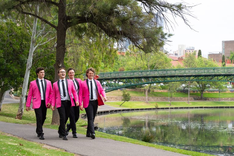 Four people in matching pink blazers walking along a riverside path beneath a bridge.