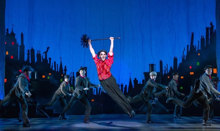 Dancer in red shirt leaps with arms raised centre-stage amid silhouetted cityscape backdrop and ensemble cast.