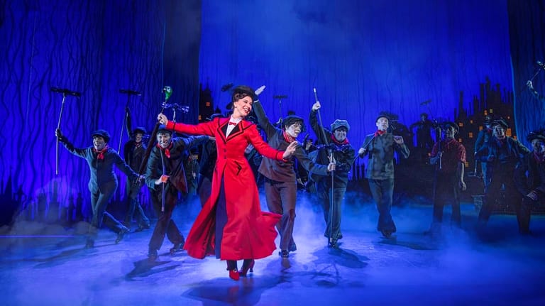 Performer in a red dress centre stage surrounded by ensemble cast under blue theatrical lighting.