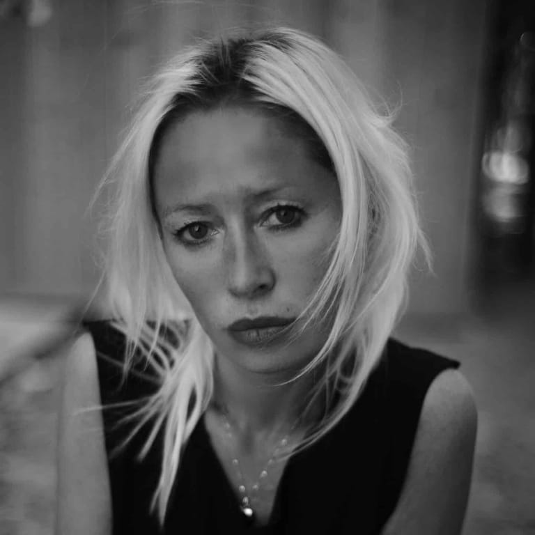 Person with blonde hair wearing a black sleeveless top and pendant necklace in a black and white portrait.