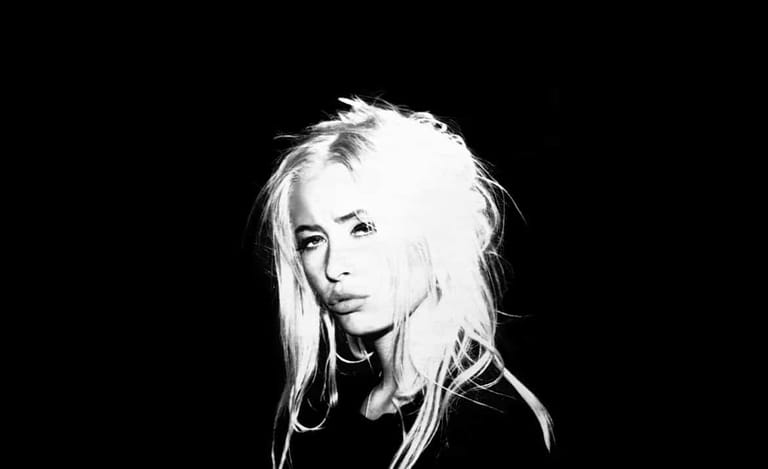 Person with long platinum blonde hair against a black background in high-contrast black and white portrait.