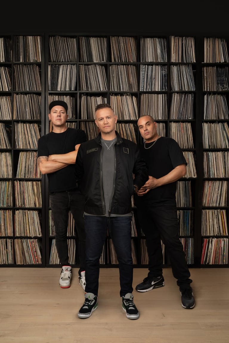 Three men in casual black clothing pose in front of a wall of stacked vinyl records.