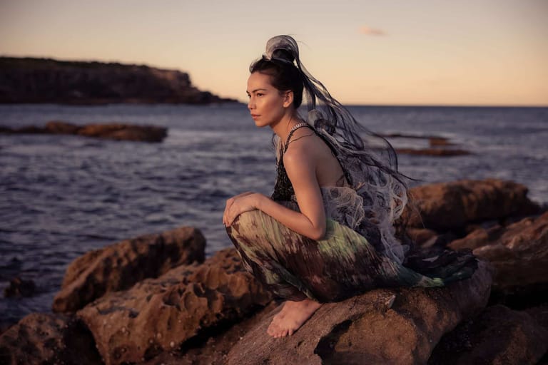 Woman with long dark hair crouched on rocky outcrop overlooking calm ocean at sunset.