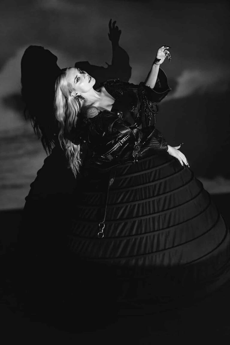 Performer in leather jacket and voluminous skirt throws head back with raised fist in dramatic black-and-white portrait.