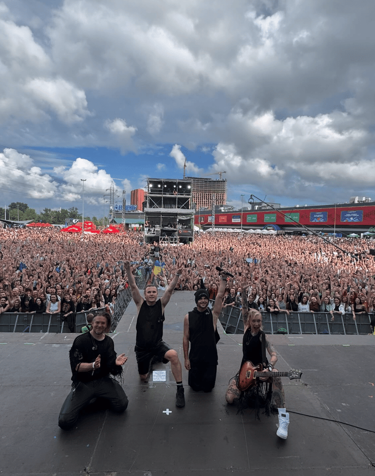 Four performers kneel on stage facing a vast outdoor festival crowd with main stage and red structures visible behind.