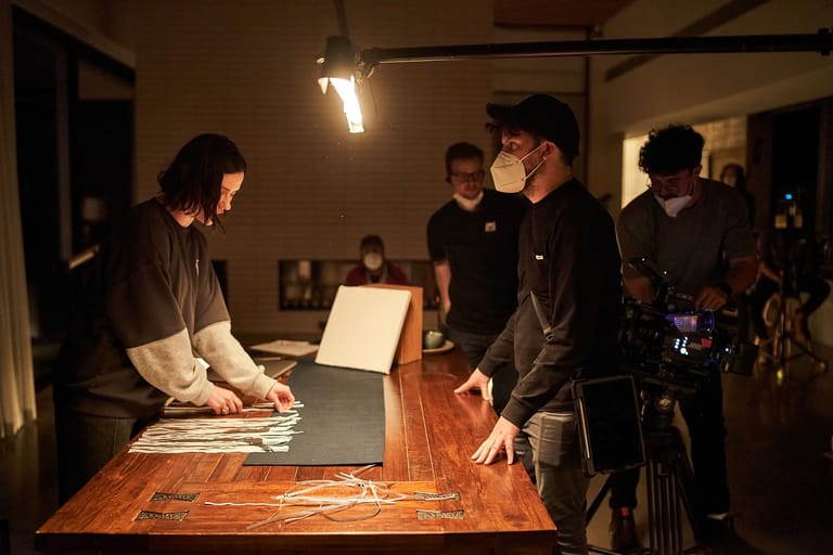 People examine artworks on a wooden table beneath warm gallery lighting during an indoor exhibition.