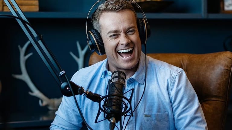 Man in light blue shirt wearing headphones and speaking into microphone in radio studio.