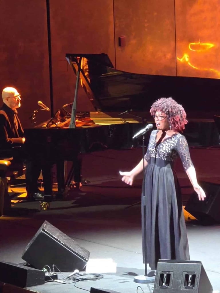 Singer in grey dress performs at microphone with pianist accompanying on grand piano, stage lit in warm amber tones.