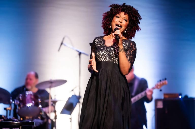 Singer in black dress with curly hair performing at microphone, drummer and guitarist visible behind on stage.