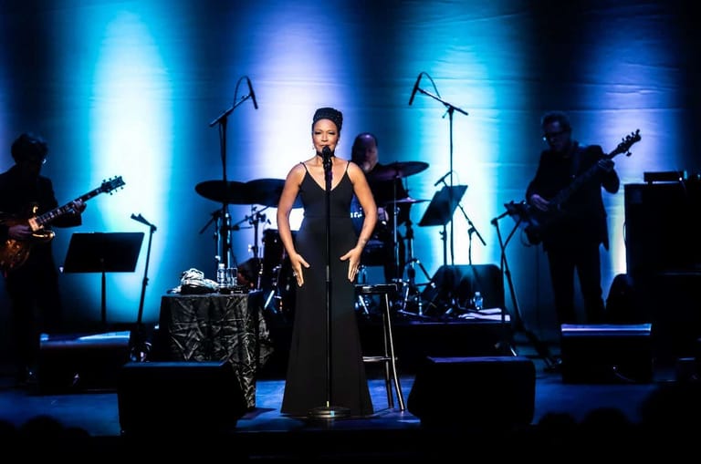 Female performer in black dress centre stage, surrounded by band members and blue stage lighting.