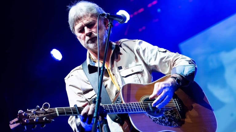 Musician with white hair playing acoustic guitar under blue stage lighting.