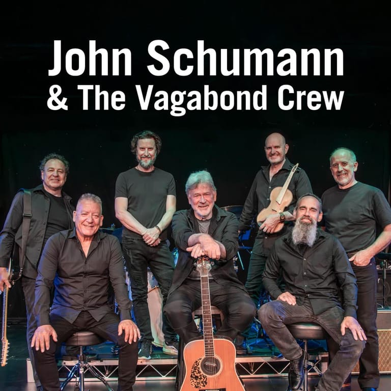 John Schumann & The Vagabond Crew band members posed together on stage with acoustic guitar.