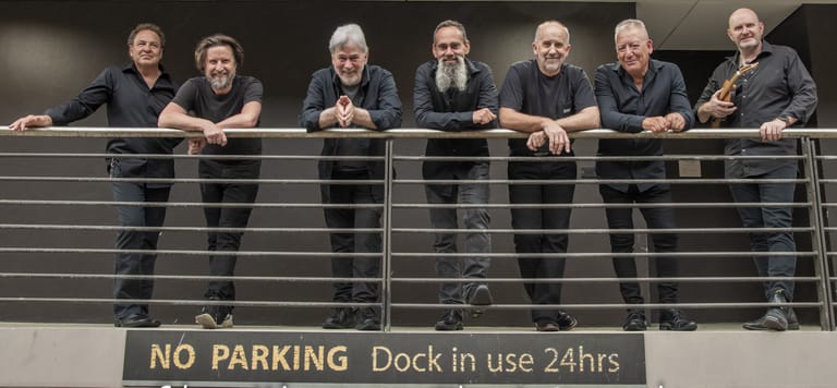 Seven men in dark suits stand together at a metal railing above a "No Parking" sign.