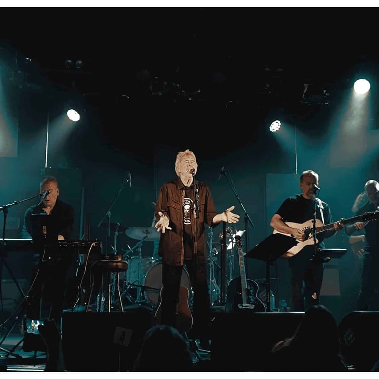 Band performs on stage under blue spotlights, with lead vocalist centre stage and musicians with instruments on either side.