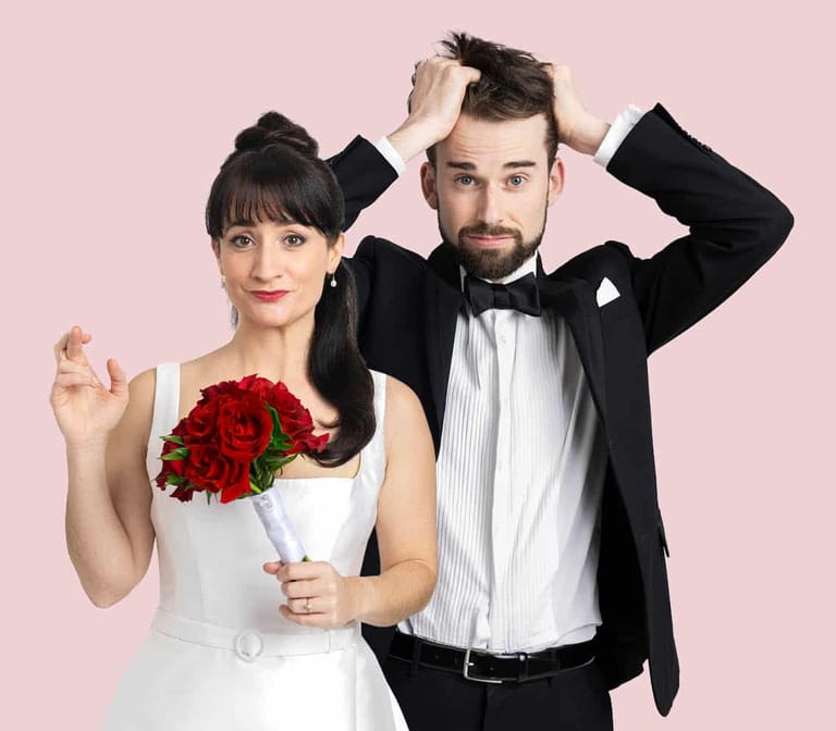 Woman in white wedding dress holding red roses stands beside man in black tuxedo against pink background.