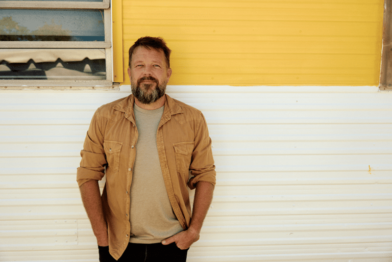 Bearded man in tan shirt standing against yellow and white corrugated wall.