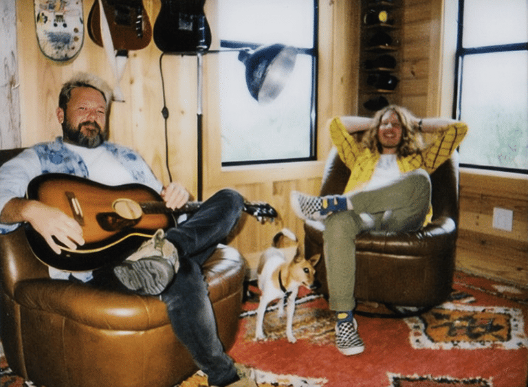 Two musicians sit in brown leather chairs in a casual studio space, one holding an acoustic guitar, the other relaxed beside him.