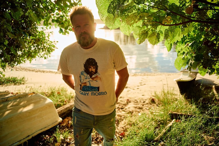 Man in printed t-shirt standing beneath dappled tree shade beside a riverbank.