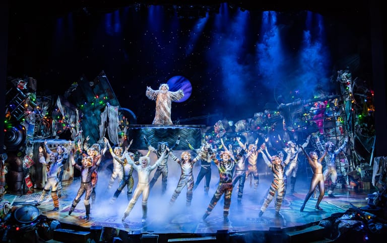 Large ensemble cast in white costumes performs on an illuminated stage under blue theatrical lighting.