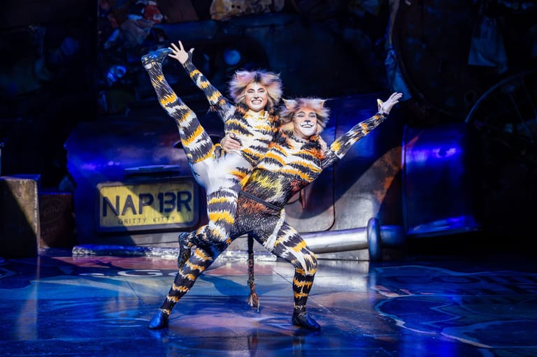 Performer in tiger costume mid-leap on stage with arms raised, illuminated by blue spotlights.